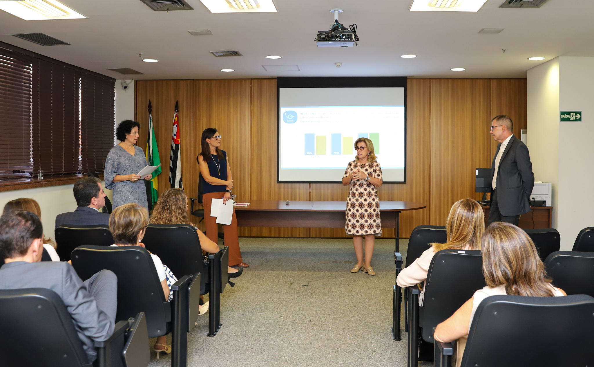 #ParaTodosVerem: três mulheres e um homem estão de pé, sendo observados por um pequeno público sentado em cadeiras pretas