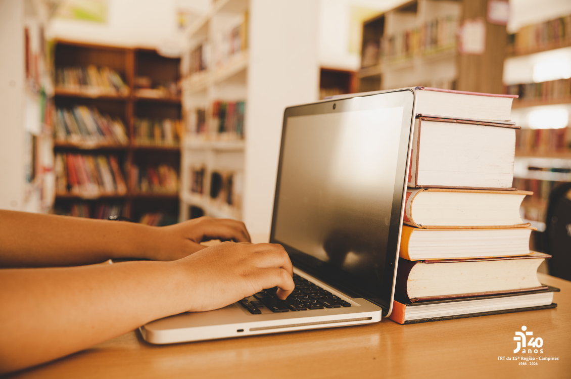 foto de mãos no teclado de um notebook sobre a mesa, escorado em pilha de livros e estantes de livros ao fundo