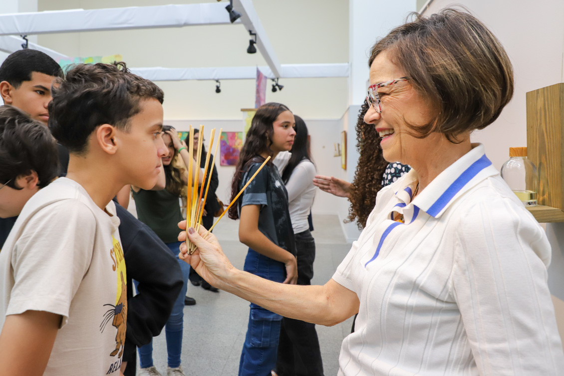 artista Michaela segura varetas de difusor para adolescente sentir o aroma. Ao fundo, outros adolescentes interagem com as obras