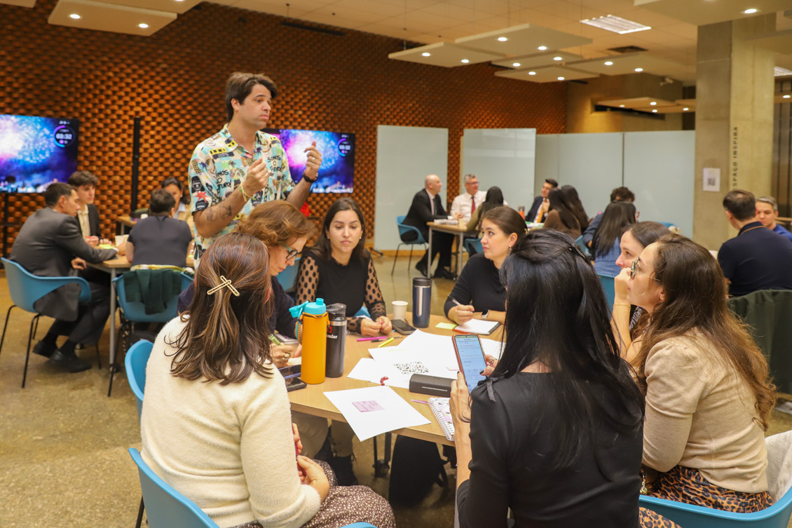 Instrutor está de pé falando com laboratoristas em atividades, agrupados em mesas redondas